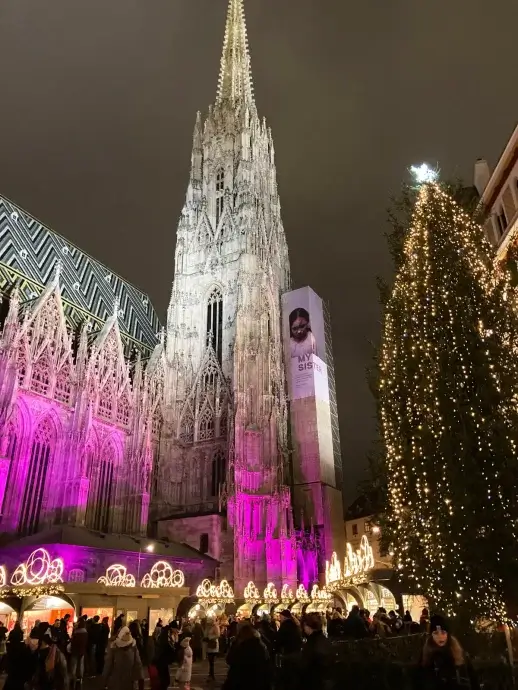 Outside of St. Stephens Cathedral in Vienna lit up with purple with stands of a Christmas Market below