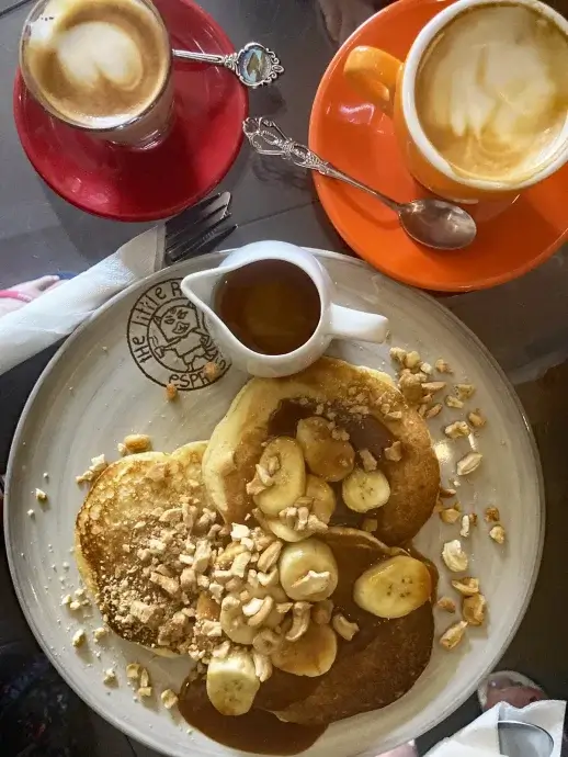 Plate with pancakes with bananas and nuts and a pitcher of syrup, two coffee cups in red and orange at the top of the image.